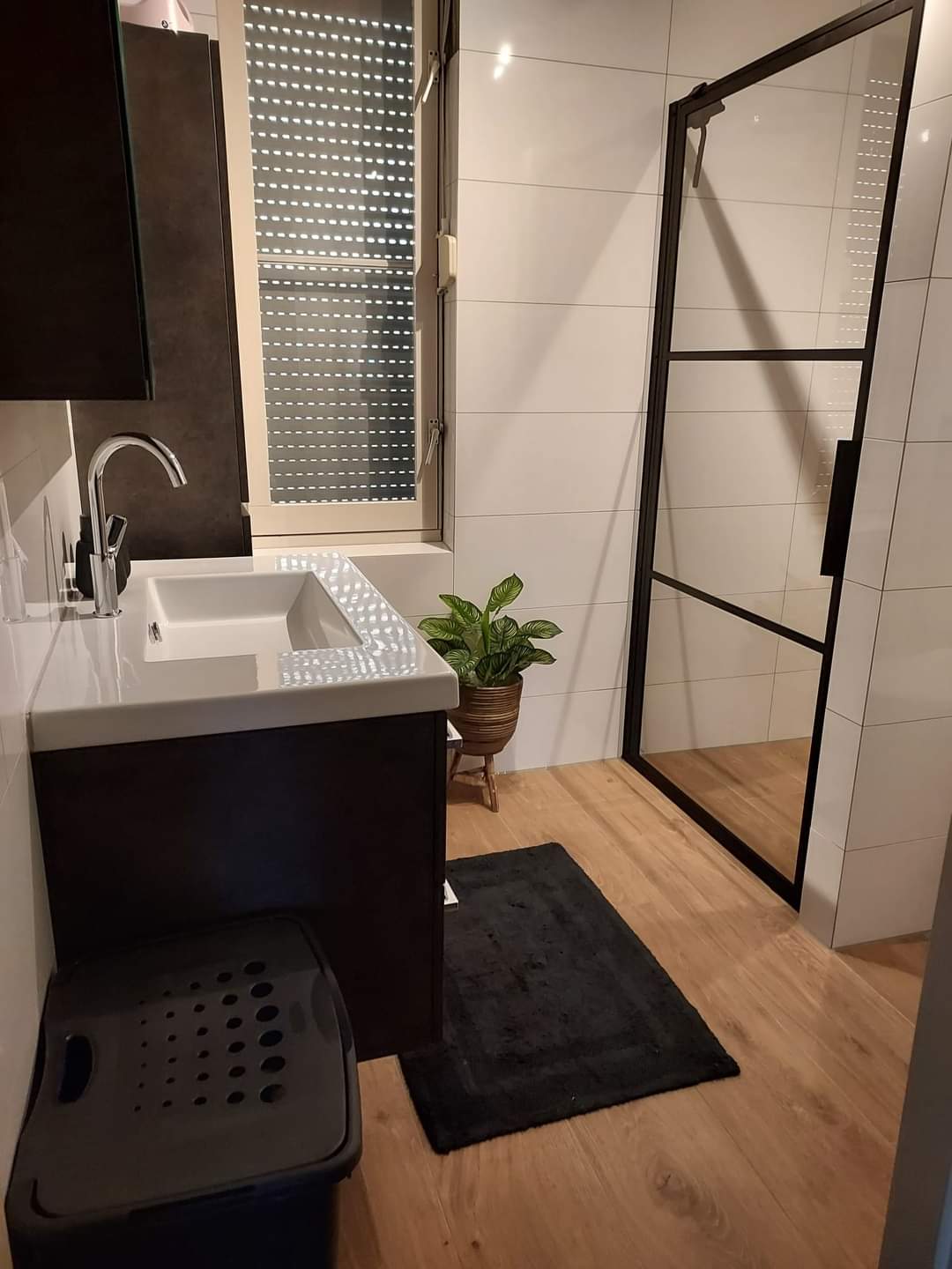 Badkamer met inloopdouche achter een zwarte stalen glaswand, witte wandtegels en een houtlook vloer